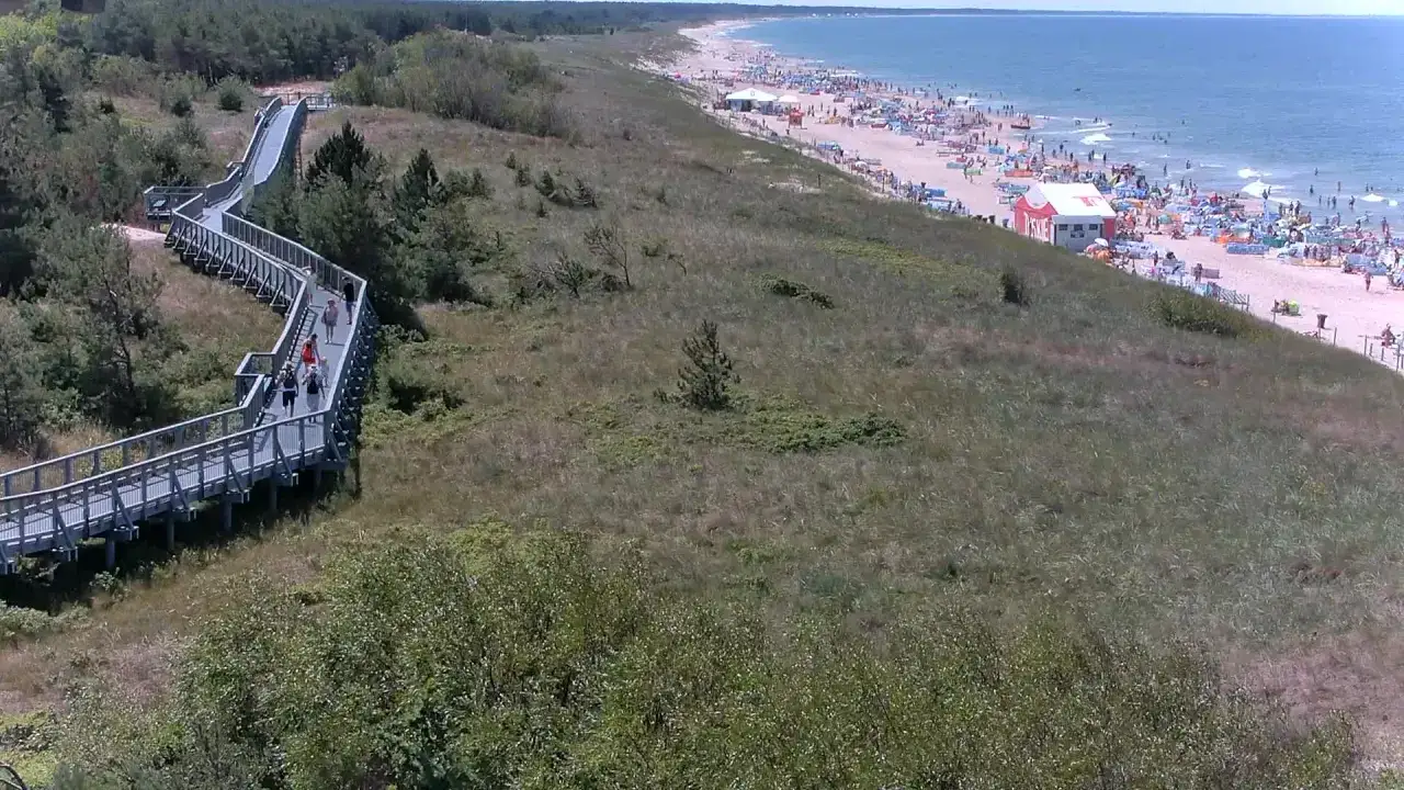 Drewniana kładka prowadzi do tętniącego życiem nadmorskiego miasta nad Wieprzą. Plaża pełna ludzi, parasoli i białego namiotu ratowniczego.