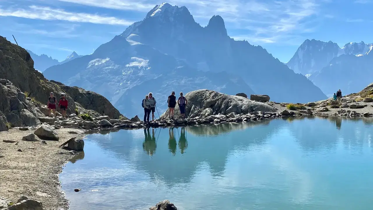 Grupa turyst&oacute;w na skalistym brzegu turkusowego jeziora, w tle majestatyczny Mont Blanc. Trasa widokowa zachwyca.