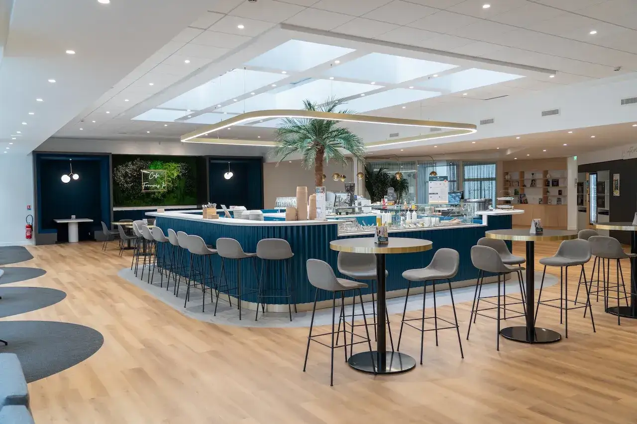Un espace restauration moderne avec un bar bleu, des tabourets gris, une plante tropicale et des tables rondes.