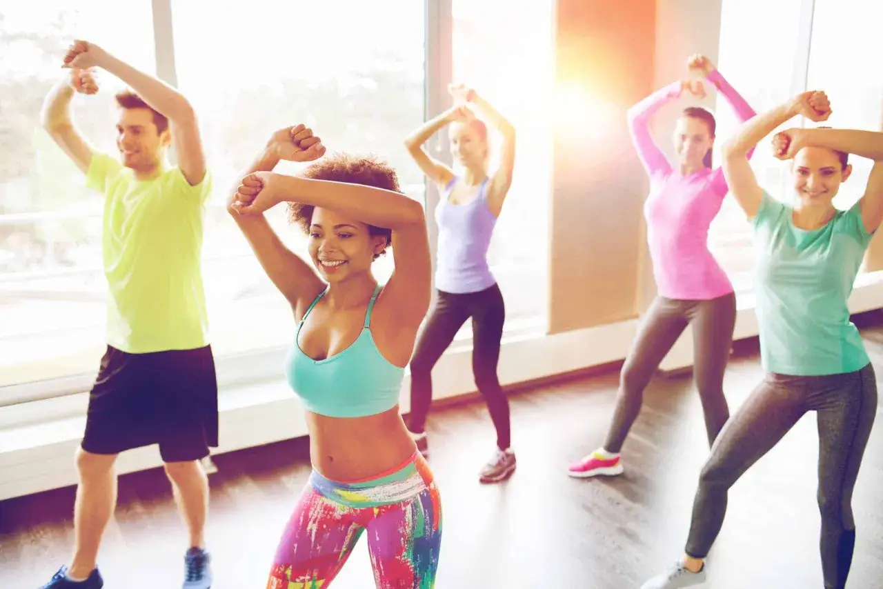 F&uuml;nf fr&ouml;hliche Menschen tanzen Zumba. Ein Mann in Gr&uuml;n und vier Frauen in Pink, T&uuml;rkis und Lila bewegen sich synchron. Ein tolles Workout f&uuml;r den Zumba Kalorienverbrauch.