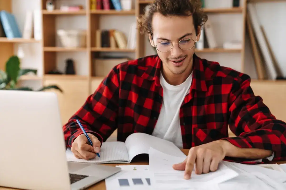 Joven con camisa a cuadros rojos y negros, frente a una laptop, escribe en un cuaderno. A pesar de que me cuesta retener informaci&oacute;n, se esfuerza por aprender.