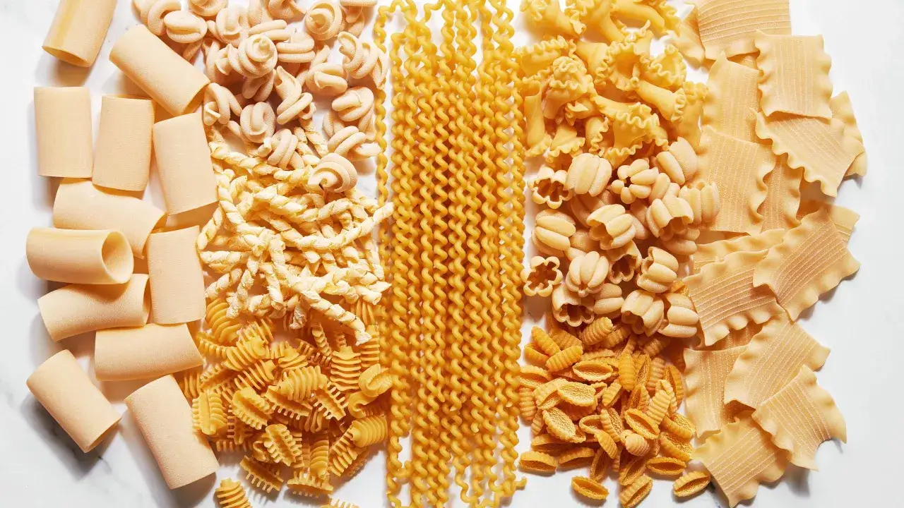 Różnorodne rodzaje makaronu ułożone na blacie. Od dużych rurek po drobne muszelki, każdy rodzaj czeka, by dowiedzieć się, jak ugotować makaron.