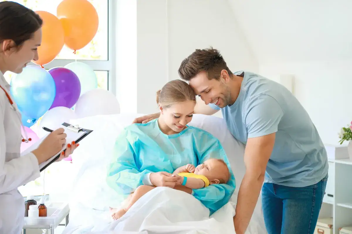 Un couple heureux avec leur nouveau-né dans une chambre d'hôpital. La maman hospitalisée tient le bébé, le papa la regarde tendrement. Des ballons décorent la pièce.