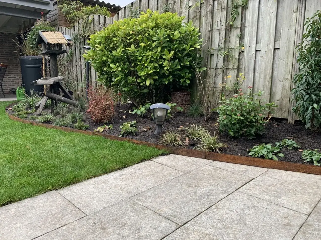 Un giardino curato con un prato verde brillante, un vialetto di lastre grigie e un'aiuola con piante e fiori. Le bordure intorno agli alberi definiscono elegantemente lo spazio.