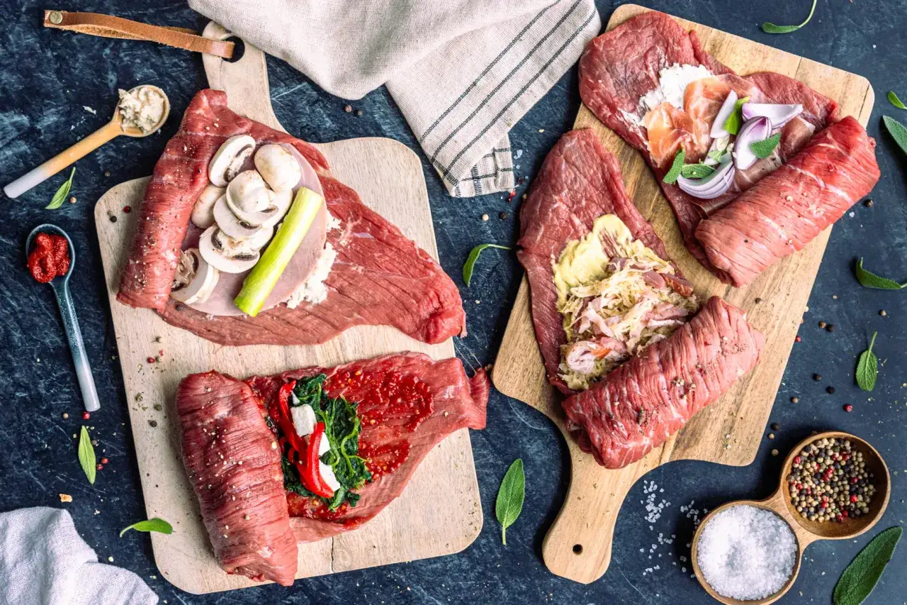 Vorbereitung für ein köstliches rouladen rezept: rohes Rindfleisch wird mit verschiedenen Zutaten wie Pilzen, Speck, Zwiebeln und Spinat belegt und auf Holzbrettern angerichtet.