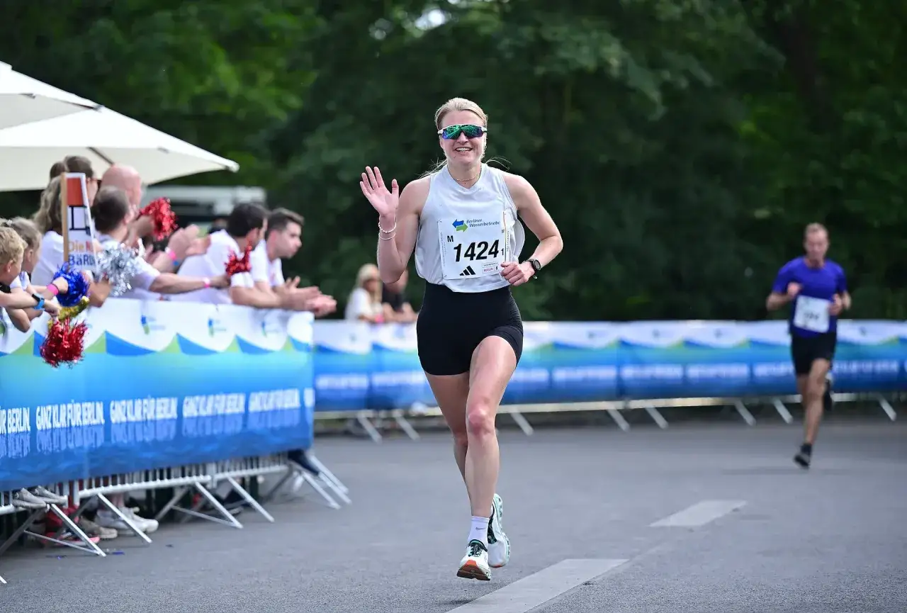 L&auml;uferin mit Startnummer 1424 winkt fr&ouml;hlich beim Berliner Teamstaffel-Lauf. Zuschauer feuern sie an.