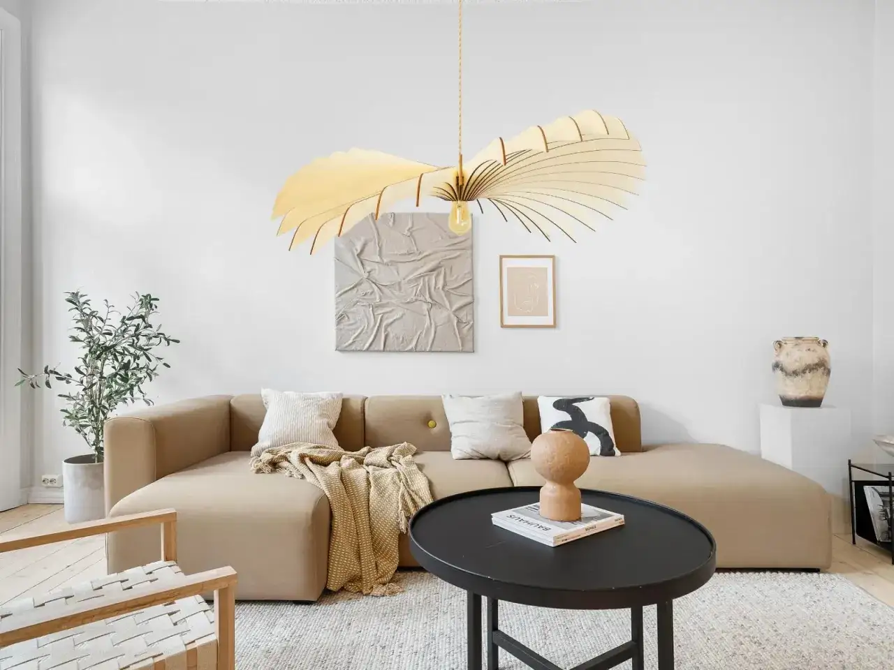 Un salon moderne avec un canap&eacute; beige, une table basse noire et une plante verte. La **tendance luminaire** est marqu&eacute;e par un lustre en bois sculptural au-dessus du canap&eacute;.