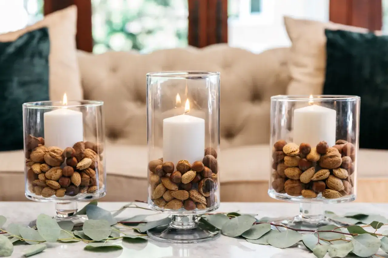 Tre candelabri fai da te con candele bianche e noci, mandorle e nocciole in vasi di vetro, decorati con rami di eucalipto.