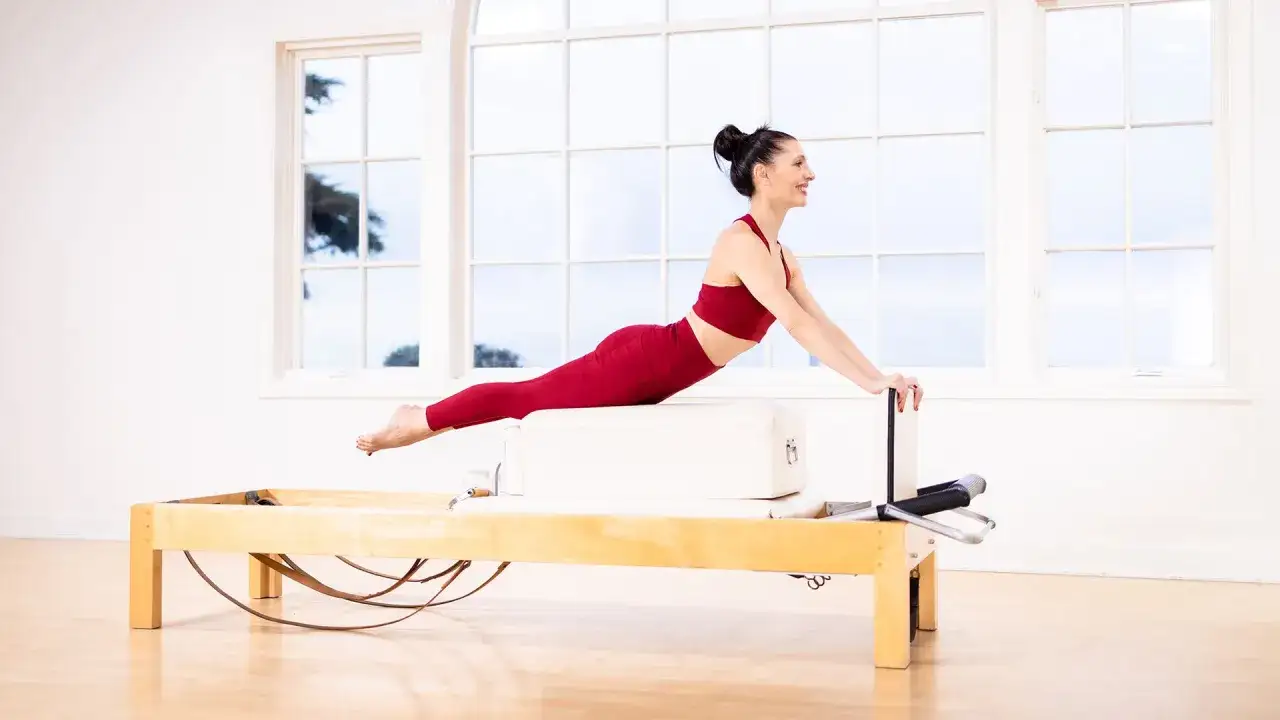 Kobieta w czerwonym stroju wykonuje ćwiczenie na reformerze, pogłębiając wygięcie kręgosłupa. To zaawansowane pilates na reformerach.