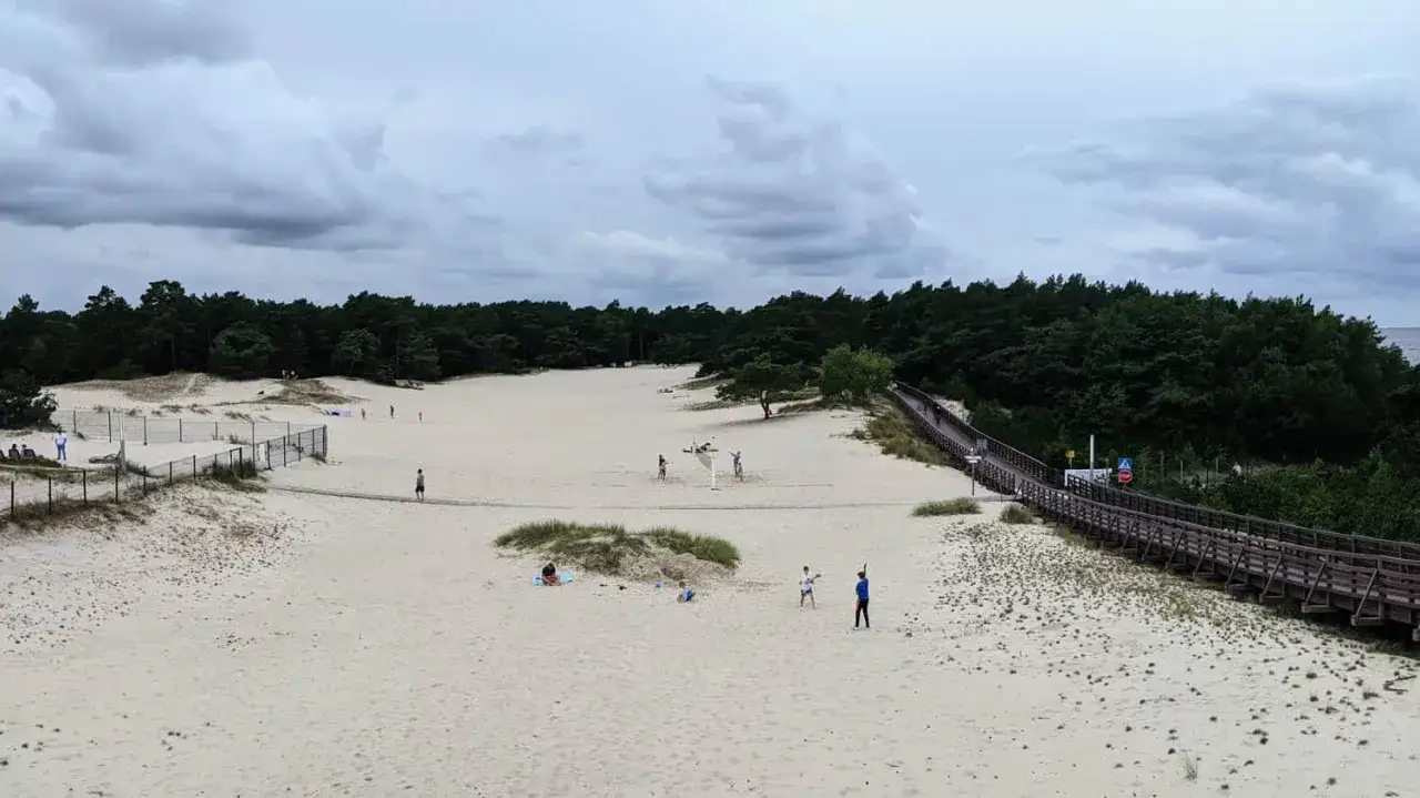 Spacer po piaszczystych wydmach w Dźwirzynie. Ludzie odpoczywają i grają w siatkówkę plażową. Drewniana kładka prowadzi przez wydmy do lasu.