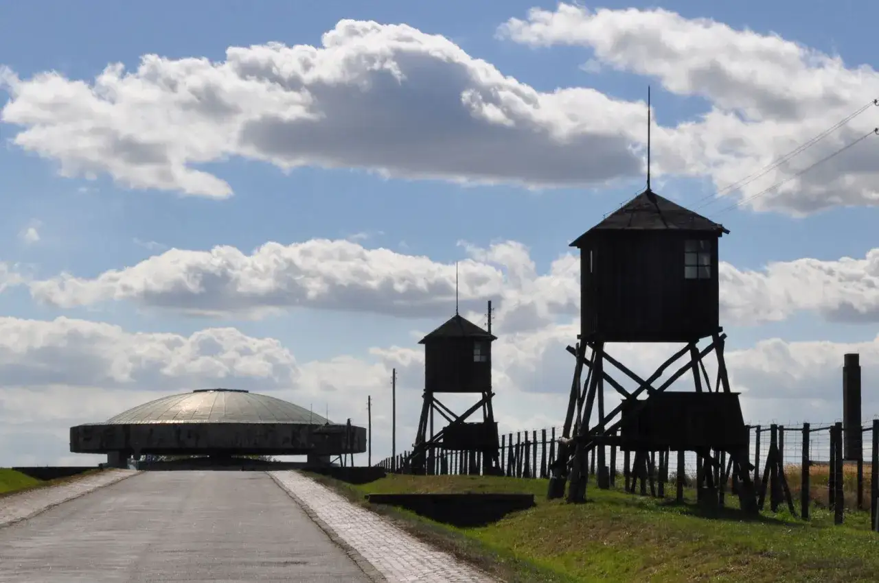 Majdanek ob&oacute;z: wieże strażnicze i pomnik w kształcie kopuły na tle błękitnego nieba z chmurami.