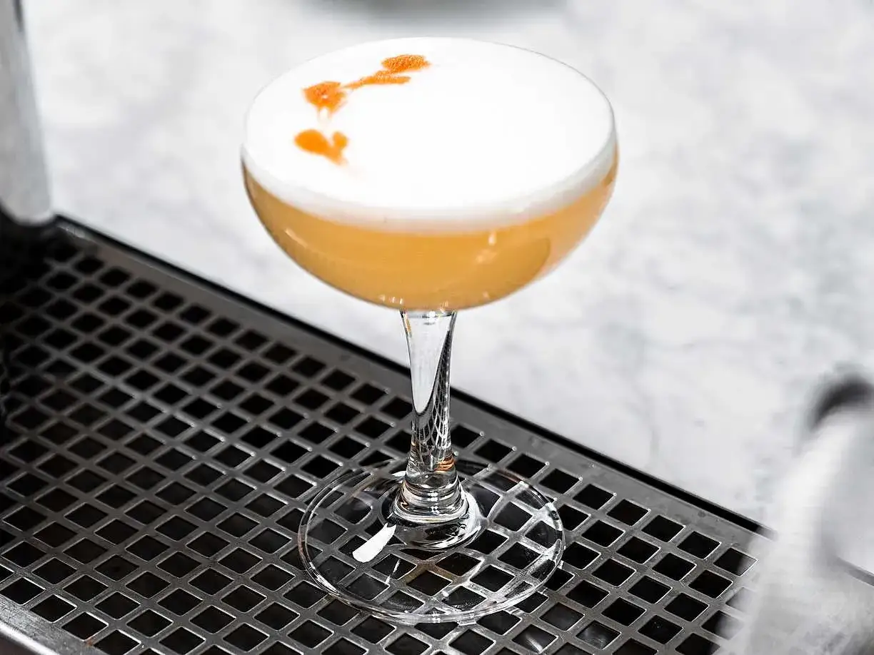 En gyllene whiskey sour med ett fluffigt skum, dekorerad med en orange virvel, står på en metallbricka.