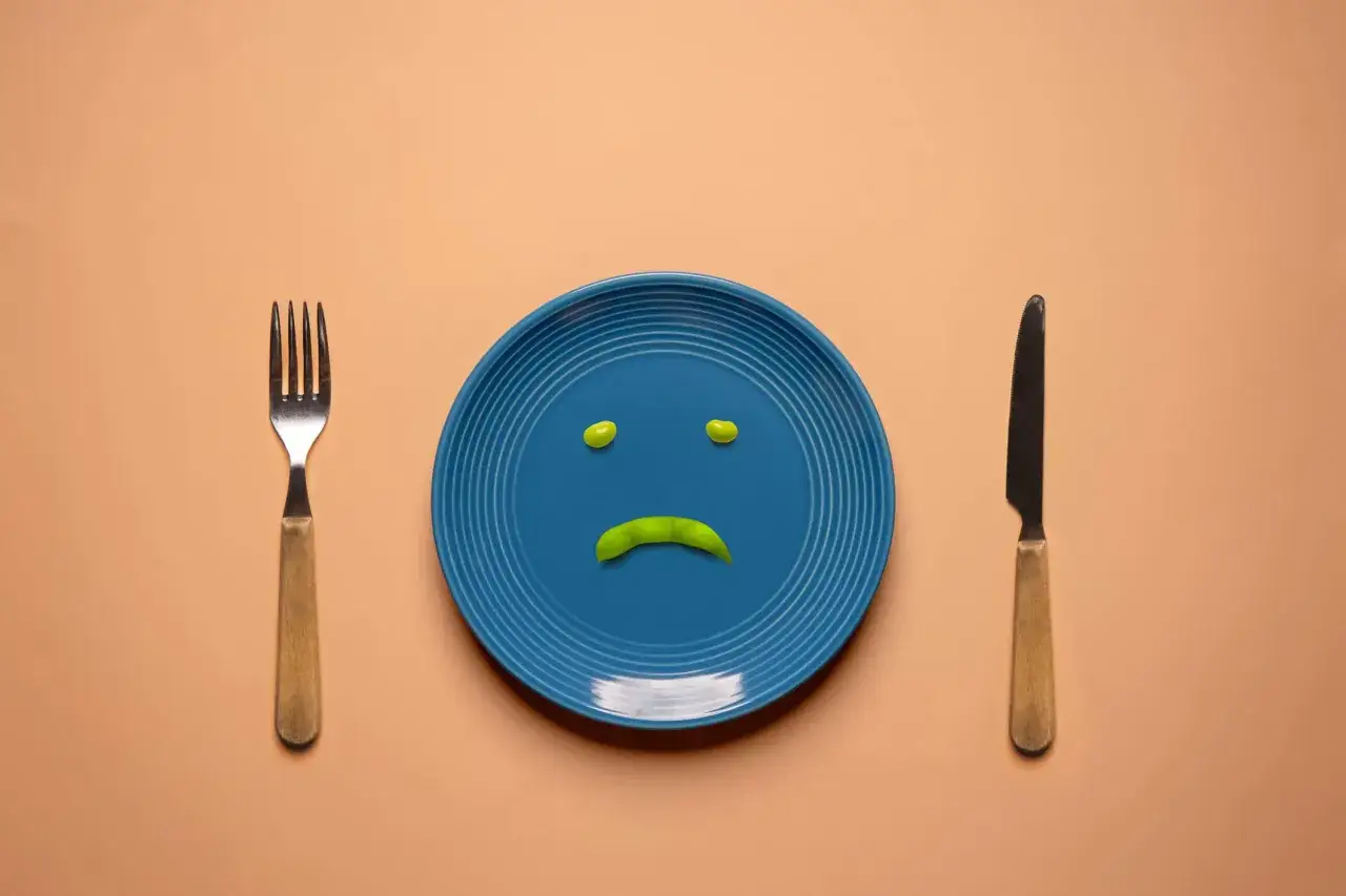 Piatto blu con faccina triste fatta di piselli, forchetta e coltello. Simboleggia i disturbi alimentari e la tristezza legata al cibo.