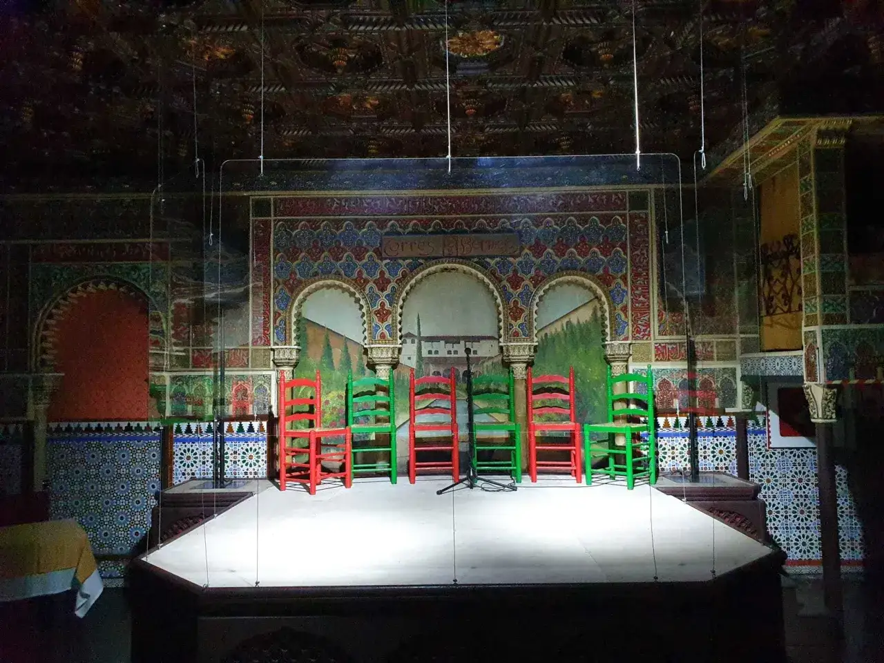 Escenario decorado con azulejos y murales, listo para un espect&aacute;culo de flamenco. Sillas rojas y verdes esperan a los artistas.