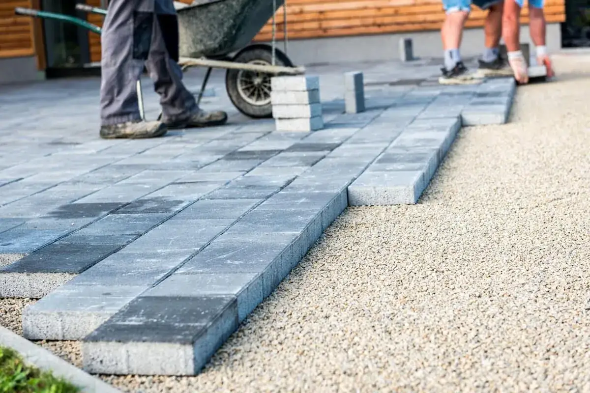 Układanie kostki brukowej na podjeździe. Pracownicy z taczką i stosem kostki przygotowują podłoże.