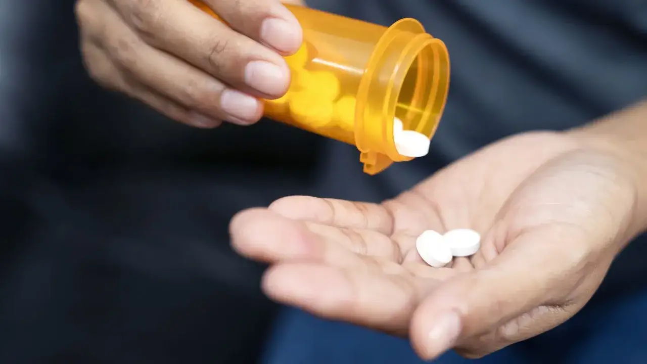 Dłoń wysypuje białe tabletki z pomarańczowej buteleczki. Wśród nich widać tabletki z napisem oxycodone.