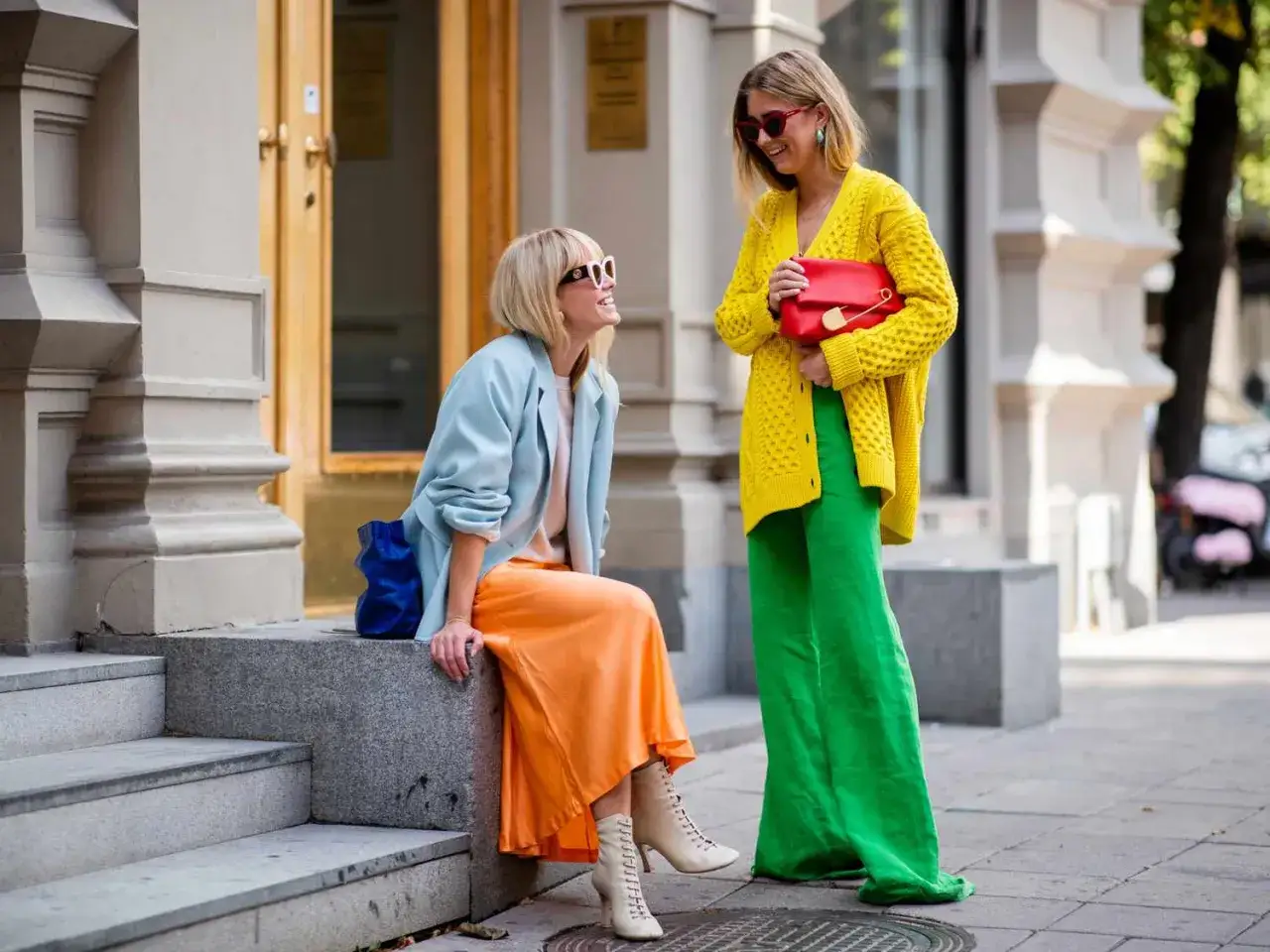 Dwie kobiety w stylizacjach color blocking: jedna w błękitnej marynarce i pomarańczowej spódnicy, druga w żółtym swetrze i zielonych spodniach.