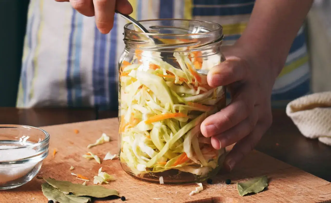 Hände packen ein Glas mit Kohl und Karotten für ein Sauerkraut Rezept. Salz, Lorbeerblätter und Pfefferkörner liegen bereit.