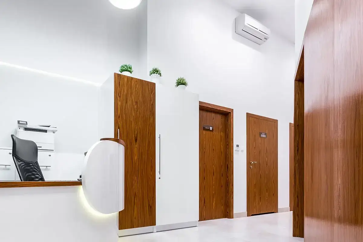 Un condizionatore d'aria a parete &egrave; installato in alto, pronto a rinfrescare due stanze. L'ambiente &egrave; moderno, con una reception e porte in legno.
