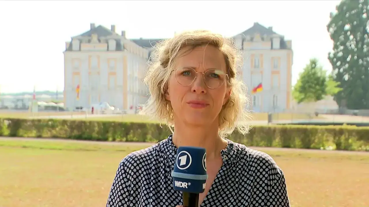 Eine Reporterin h&auml;lt ein Mikrofon vor einem Schloss, auf dem die Flaggen von Deutschland und Frankreich wehen.