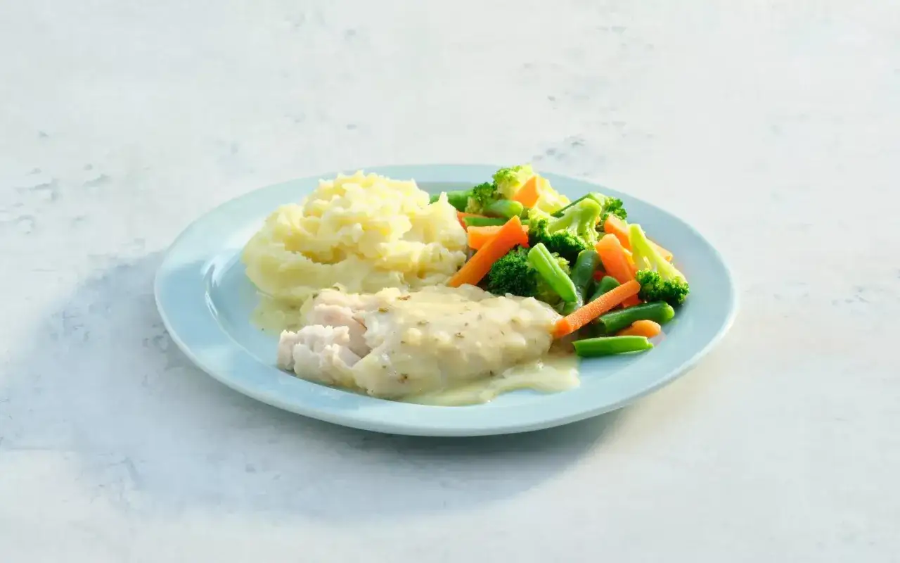 Plato azul con pur&eacute; de patatas cremoso, pescado blanco en salsa y verduras al vapor como br&oacute;coli, zanahoria y jud&iacute;as verdes.