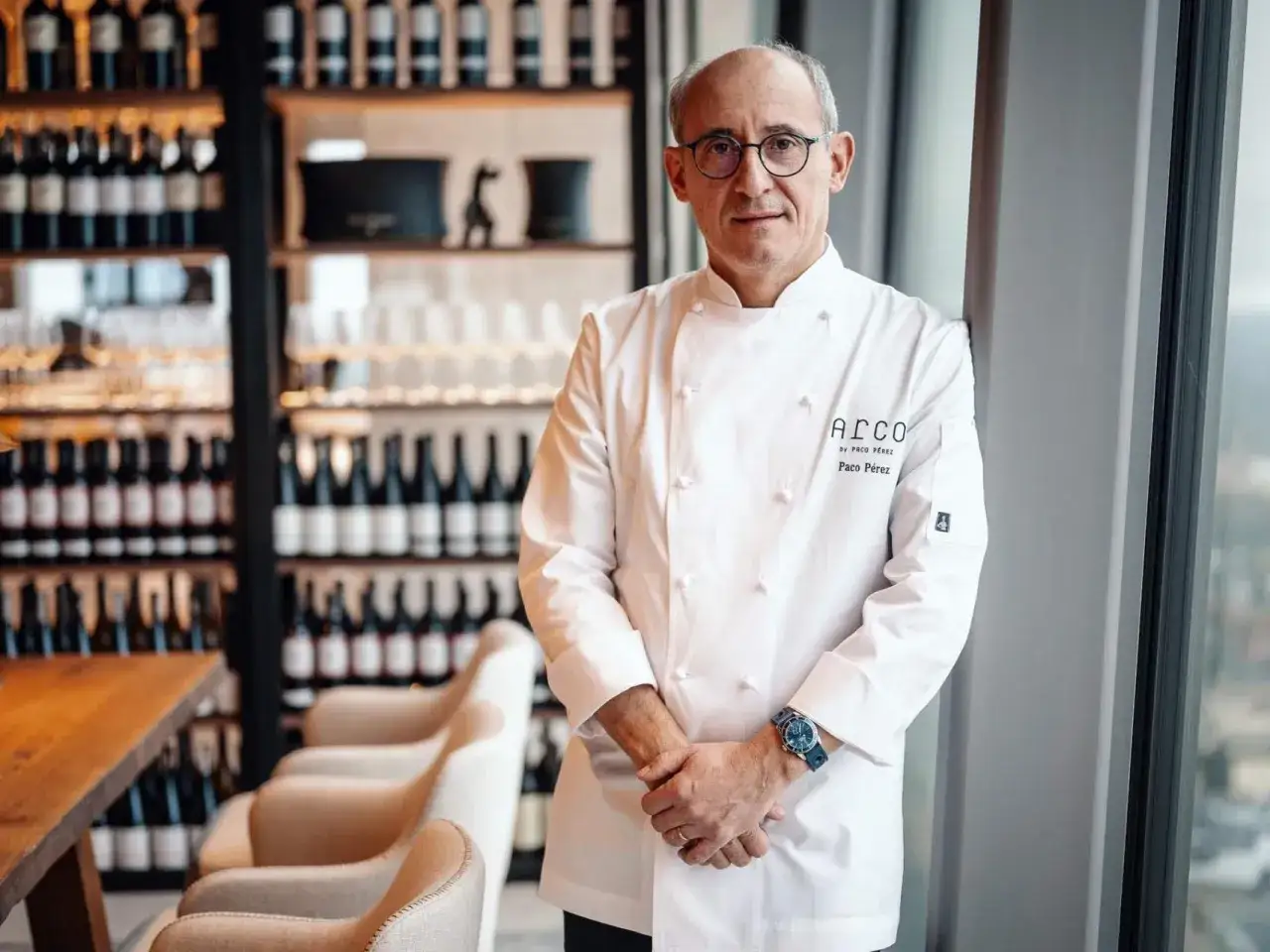 Chef Paco Pérez, con gafas y chaquetilla blanca, posa en un restaurante con estanterías de vino.