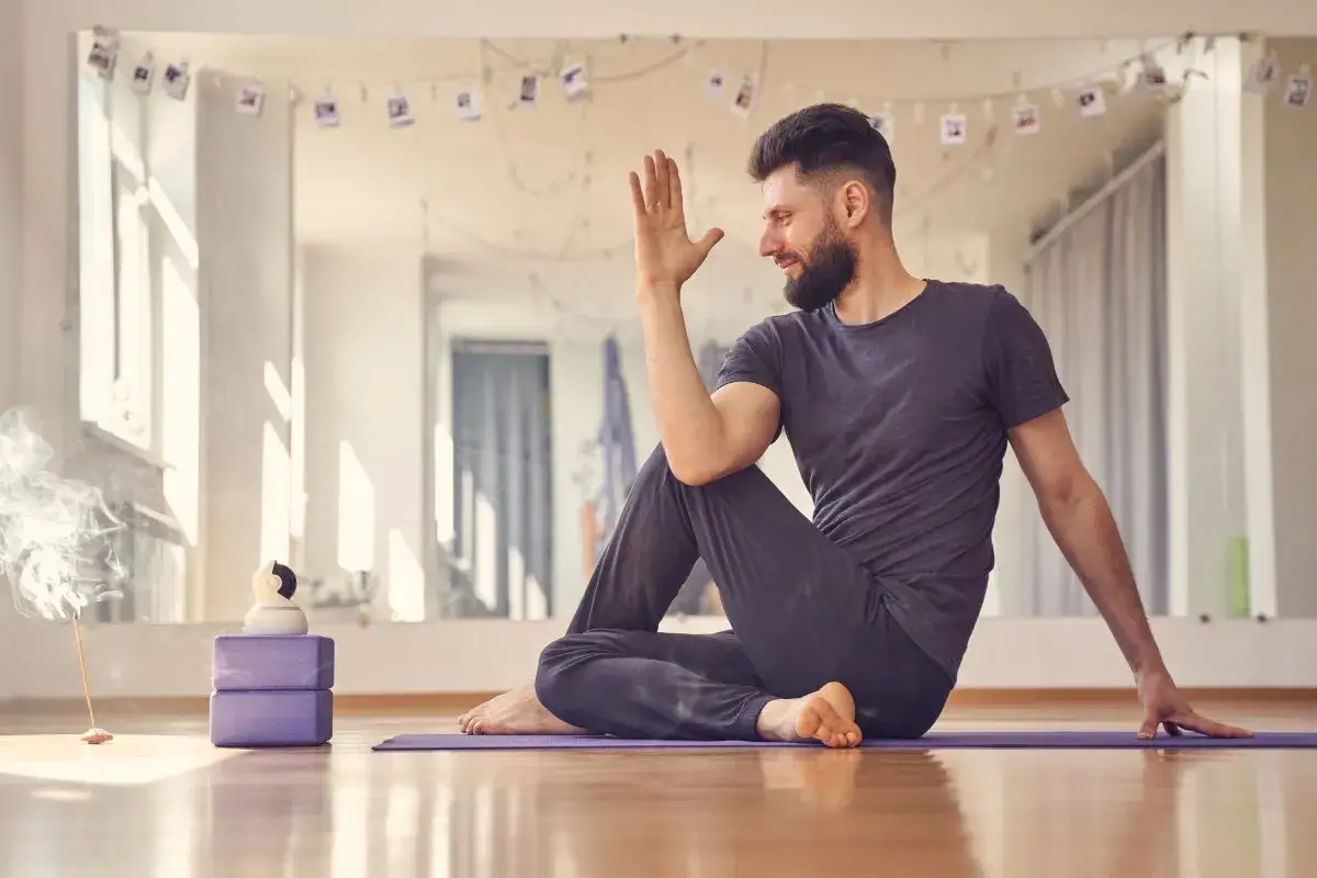 Hombre sonriente practicando yoga para hombres en un estudio luminoso. Postura de torsión sentada, con incienso y bloques de yoga.