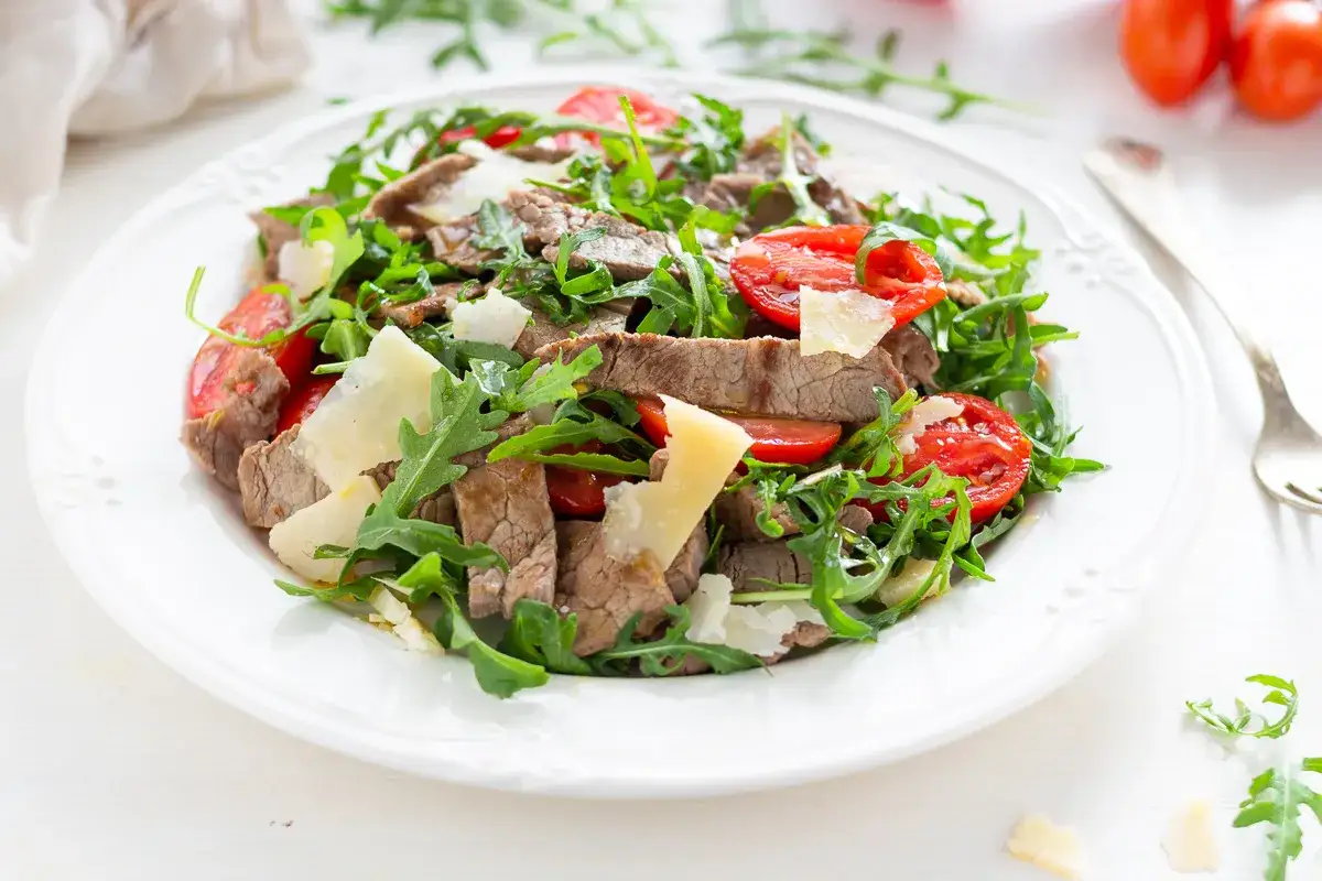 Deliziosi straccetti di manzo con pomodorini freschi, rucola croccante e scaglie di Parmigiano Reggiano su un piatto bianco.