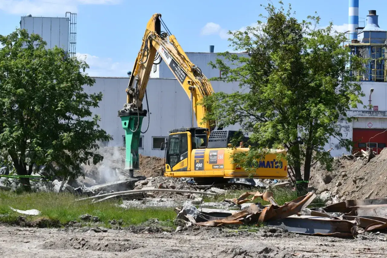 Żółta koparka z młotem hydraulicznym prowadzi rozbiórkę gruzowiska. W tle budynek przemysłowy i drzewa.