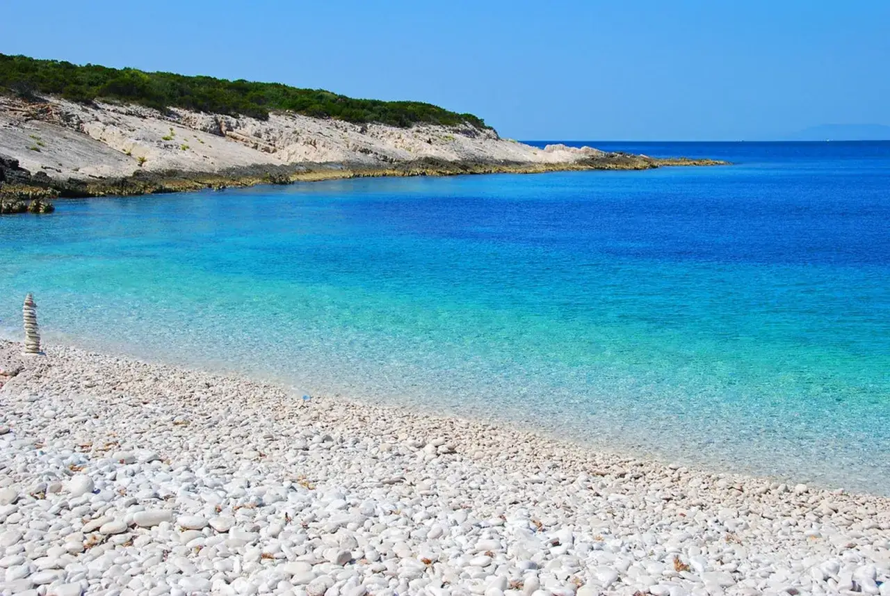 Spiaggia di ciottoli bianchi e mare cristallino, un angolo di **Croazia mare pi&ugrave; bello** con rocce e vegetazione mediterranea.