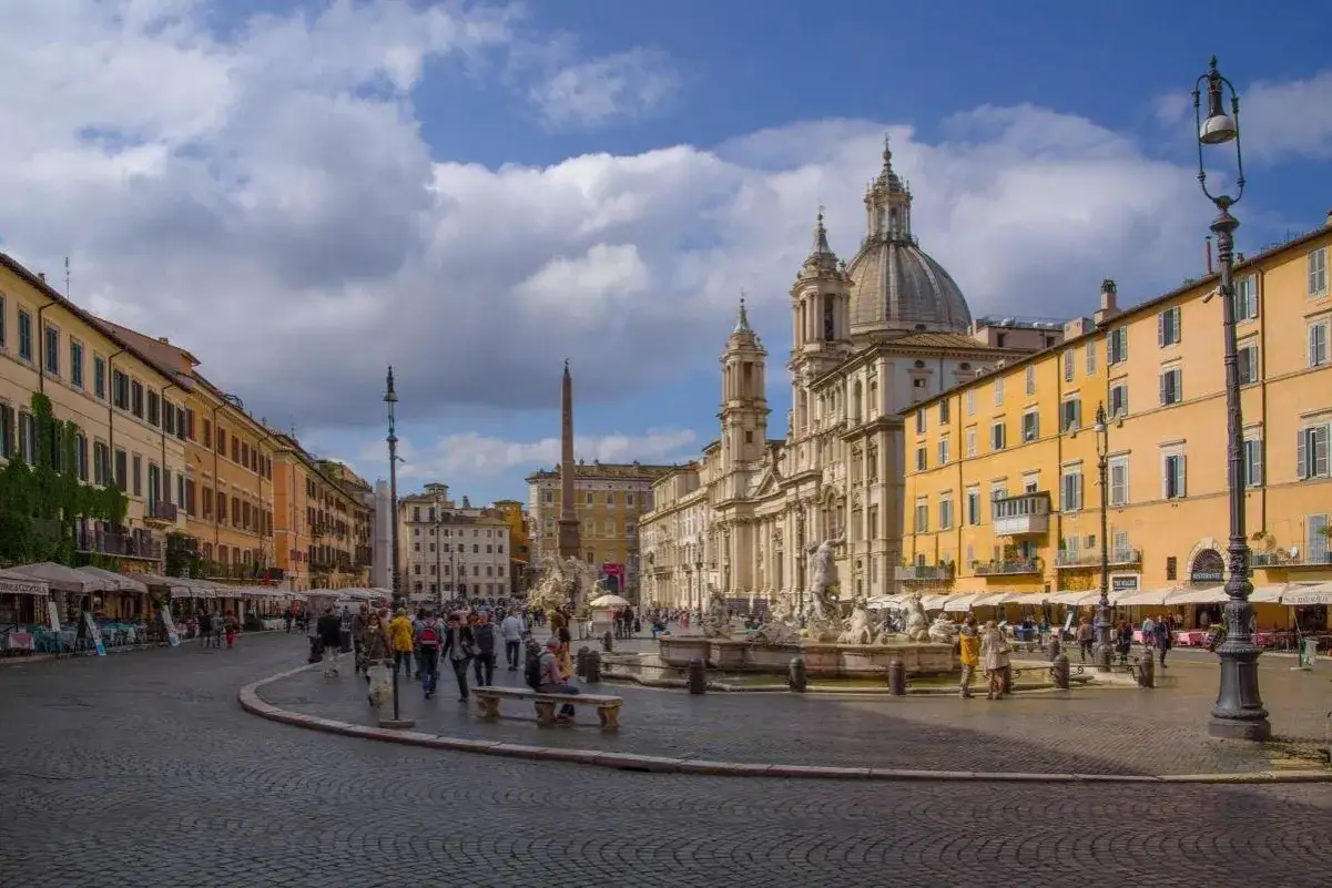 Tętniąca życiem Piazza Navona w Rzymie, z Fontanną Czterech Rzek i obeliskiem w tle. Ludzie spacerują, siedzą na ławkach, ciesząc się atmosferą.