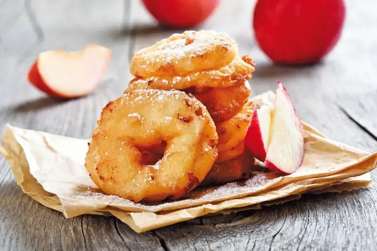 D&eacute;licieux Apfelkiechle, beignets de pommes saupoudr&eacute;s de sucre glace, sur du papier brun, avec des quartiers de pommes rouges en arri&egrave;re-plan.