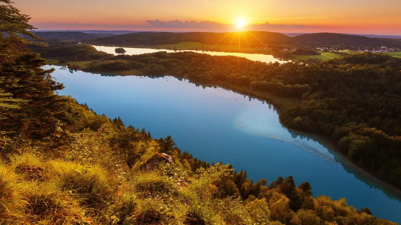 Vue panoramique d'un lac bleu intense au coucher du soleil, parfait pour une randonnée Jura 2 jours. Forêts verdoyantes et collines en arrière-plan.