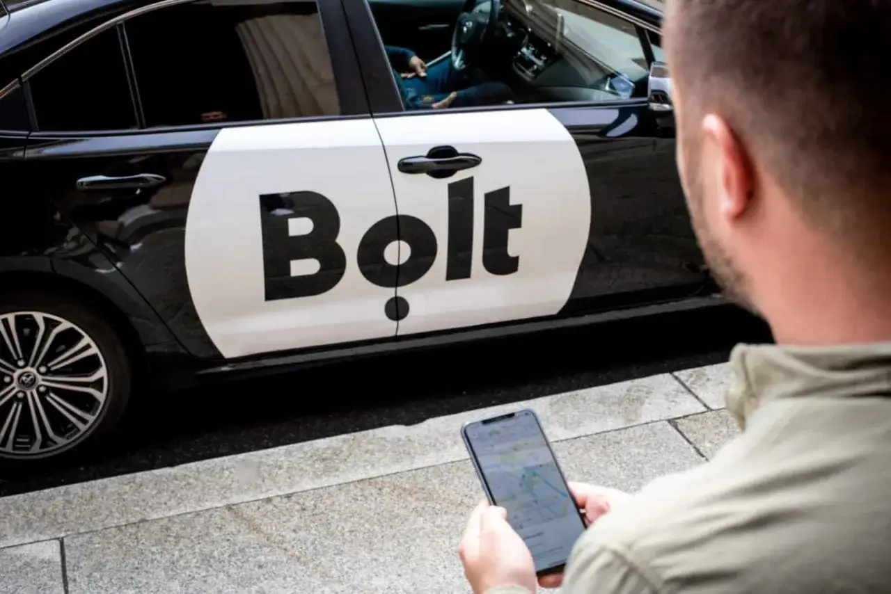 Ein Mann mit Smartphone wartet auf seinen Bolt Fahrer neben einem Auto mit Bolt Logo.