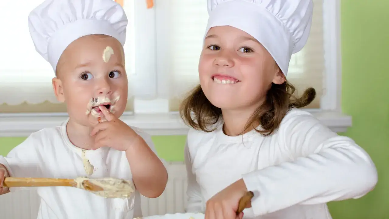 Zwei Kinder in Kochm&uuml;tzen, eines mit Mehl im Gesicht, das andere l&auml;chelnd. Das kleine Kind probiert den Teig, als ob es **Mehl roh essen** w&uuml;rde.