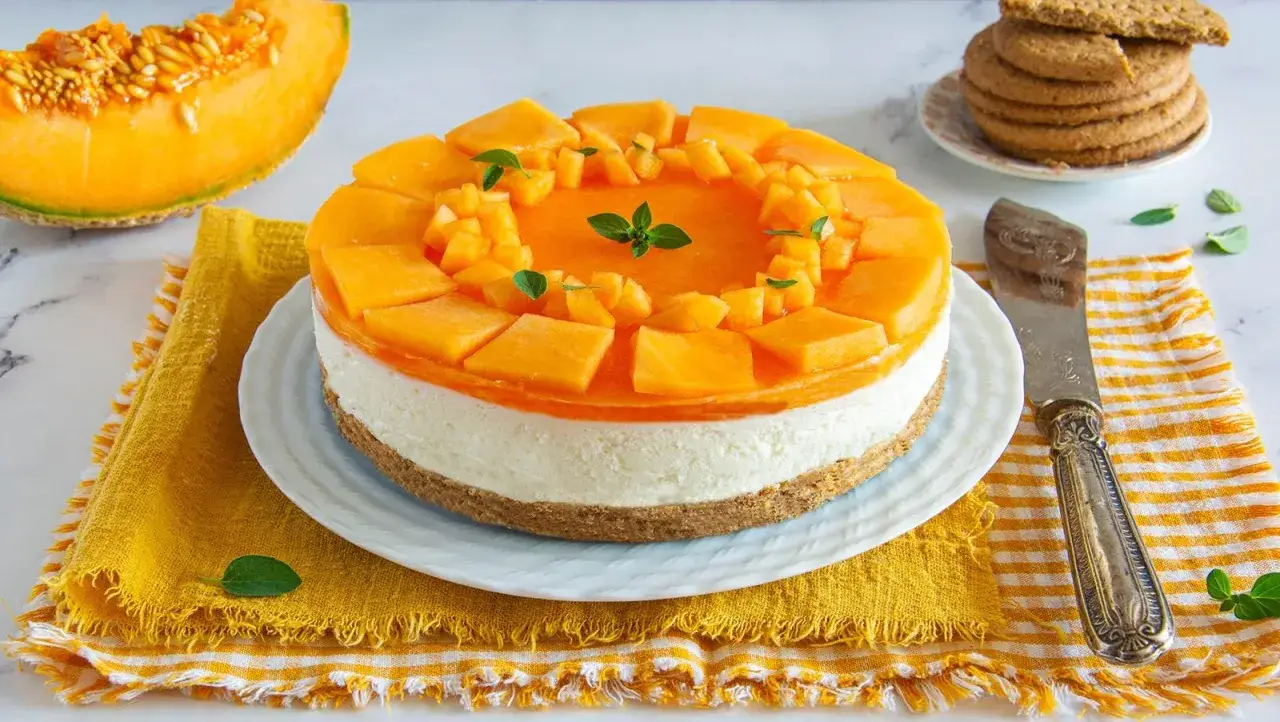 Deliziosa cheesecake al melone con cubetti di frutta fresca e gelatina arancione, su una base croccante. Accanto, una fetta di melone e biscotti.