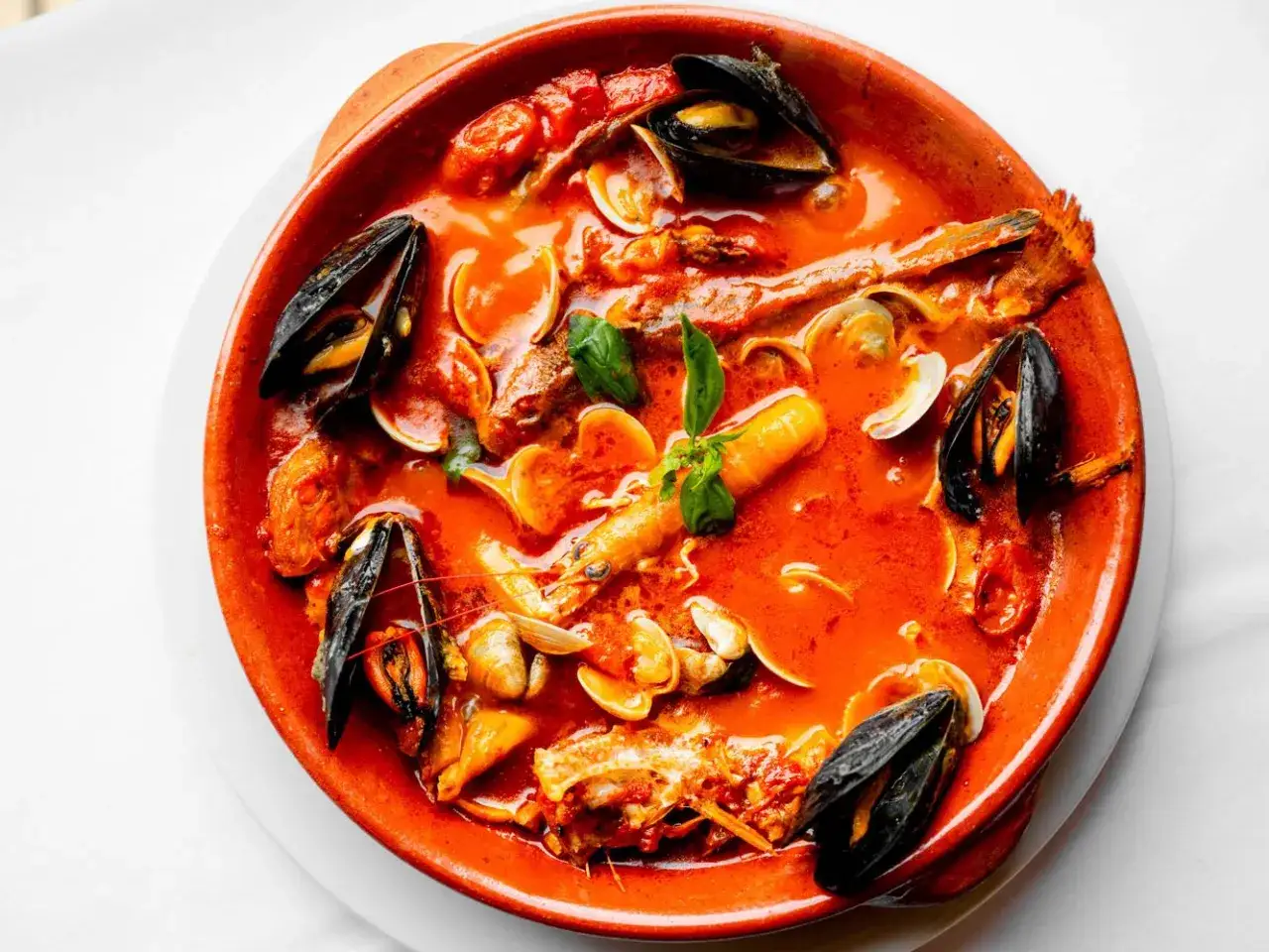 Una ricca zuppa di pesce in una ciotola di terracotta, con cozze, vongole e gamberi in un brodo rosso.