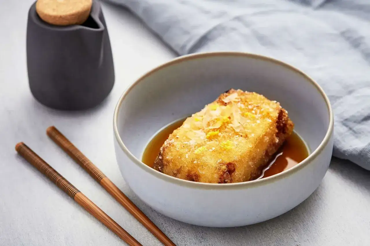 A golden, crispy aburaage tofu sits in a savory broth, topped with delicate flakes and zest. Chopsticks and a small pitcher complete the elegant presentation.