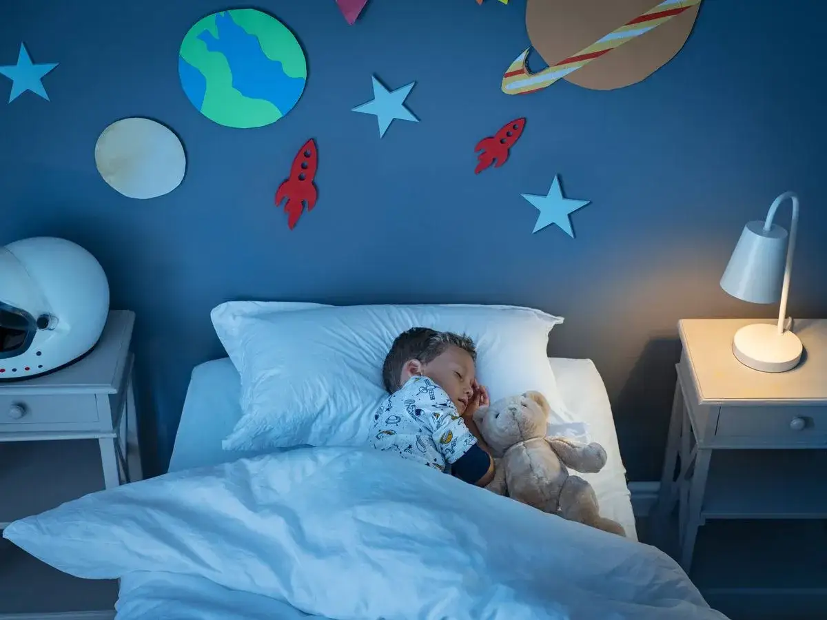 Niño duerme solo en su cama, rodeado de decoración espacial. Un peluche lo acompaña, un ejemplo de como hacer que un niño duerma solo.