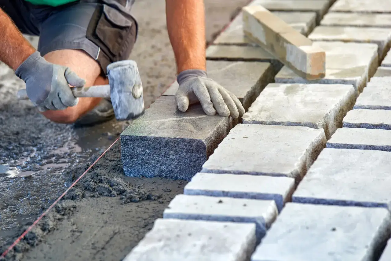 Układanie kostki brukowej na podsypce. Widać dłoń w rękawicy i młotek gumowy, a także przygotowany suchy beton proporcje.
