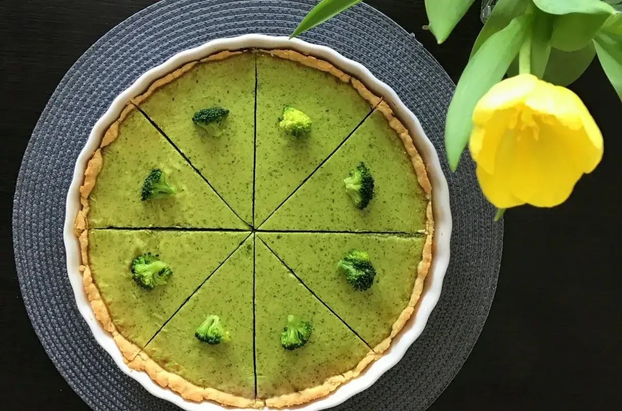 Zielona tarta z brokułami, pokrojona na 8 kawałk&oacute;w, ozdobiona r&oacute;życzkami brokuł&oacute;w. Obok ż&oacute;łty tulipan.
