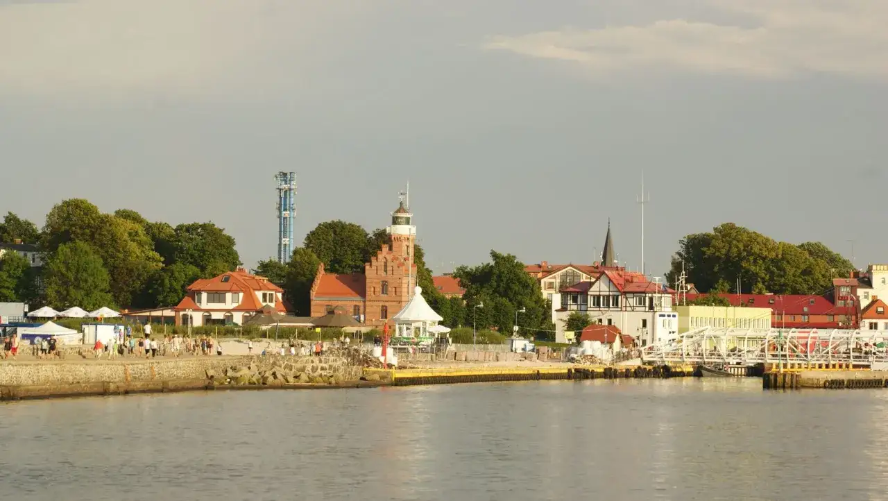 Widok na port z charakterystyczną ceglaną latarnią morską w Ustce. Spacerowicze na nabrzeżu, biały most i zielone drzewa.