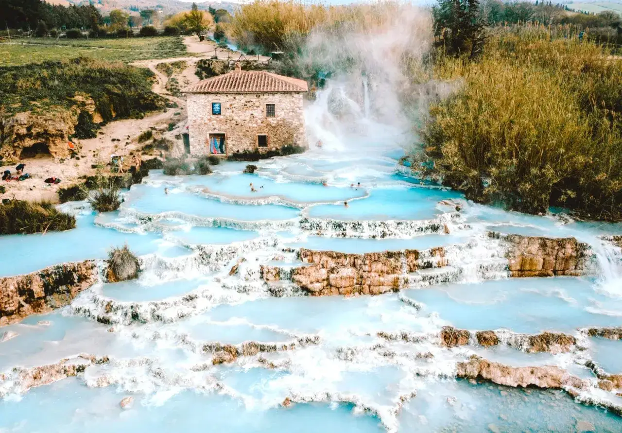 Nat&uuml;rliche t&uuml;rkisfarbene Becken und Wasserf&auml;lle bei Bagni di San Filippo, mit einem alten Steinhaus im Hintergrund.
