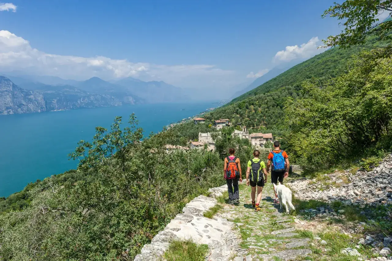 Tre escursionisti con zaini e un cane camminano su un sentiero di pietra con vista sul Lago di Garda. Perfette unternehmungen gardasee!