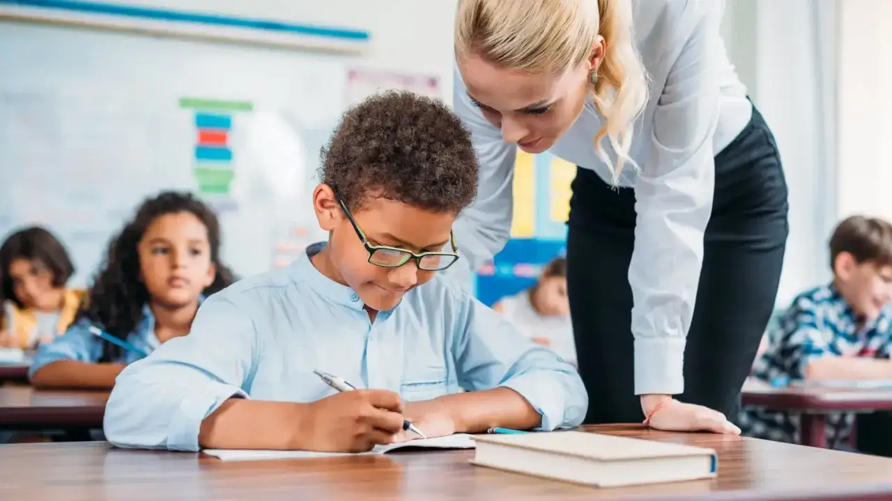 Maestra ayuda a un niño con gafas a escribir, mostrando apoyo ante un posible trastorno específico del aprendizaje.