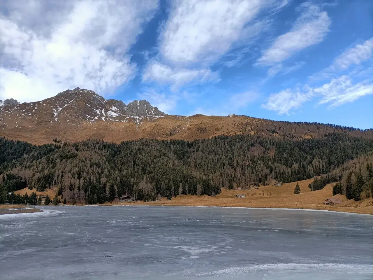 Il lago di Lova ghiacciato riflette il cielo azzurro con nuvole bianche. Montagne innevate e boschi scuri incorniciano il paesaggio.