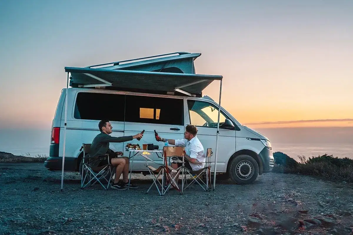 Zwei Männer stoßen mit Bier an, während sie am Abend vor ihrem Campervan sitzen. Ein perfekter Moment für Barcelona Highlights.