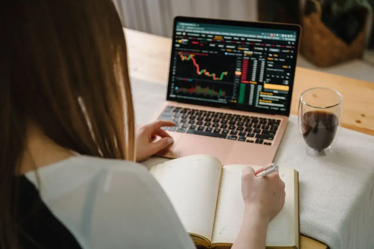 Donna studia grafici finanziari sul laptop, prendendo appunti per il suo trading online. Un caff&egrave; la accompagna.