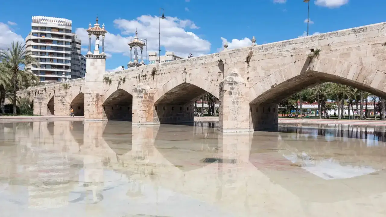 El **puente de la Exposici&oacute;n**, con sus arcos de piedra, se refleja en el agua serena, bajo un cielo azul con nubes.