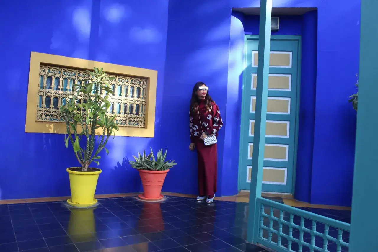 Donna con occhiali a cuore posa davanti al blu intenso del museo Yves Saint Laurent Marrakech, con piante in vaso e una porta turchese.